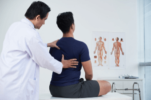 Doctor examining a male patient’s back and shoulders in a medical office with anatomical charts on the wall.