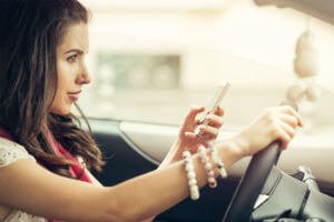 Woman sitting in a car holding a smartphone in one hand and steering the wheel with the other.