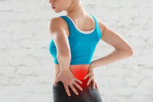 Woman in blue tank top holding her lower back, indicating pain or discomfort with a red highlight on the affected area.