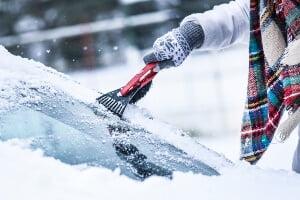 a driver wrapped up for winter using an ice scraper to clear a windshield