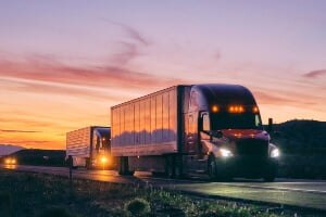 two semi-trucks driving at dusk with headlights of cars behind them