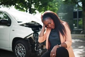 young black woman holding her head and kneeling by her damaged car