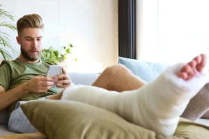 A young white man with blond hair and a cast on his leg sitting on a sofa looking at his phone