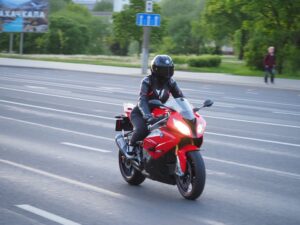 Motorcyclist in black gear riding a red sports bike on a multi-lane road with trees and pedestrians in the background.