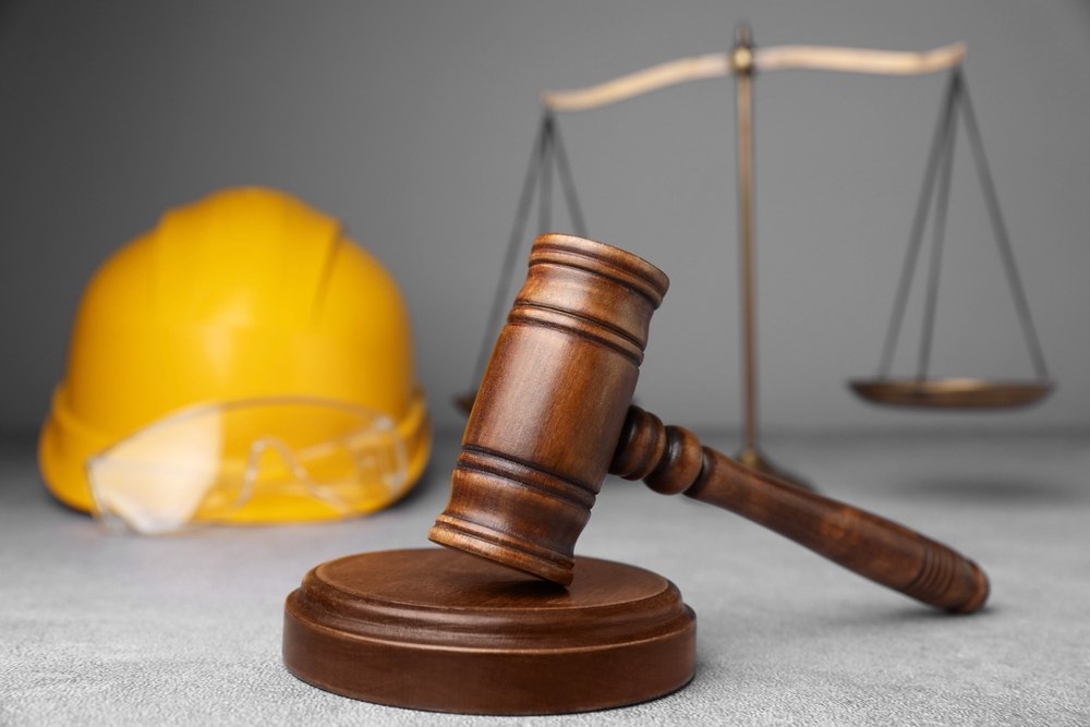 Wooden judge gavel in front of a yellow construction helmet, safety glasses, and a blurred balance scale on a gray surface