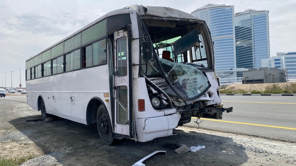Damaged white bus with broken windshield and front, parked on roadside near modern glass buildings under a cloudy sky